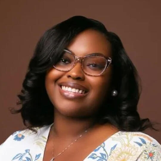 A headshot of a dark skinned Black woman smiling, wearing glasses and a bright smile, with wavy shoulder length hair.